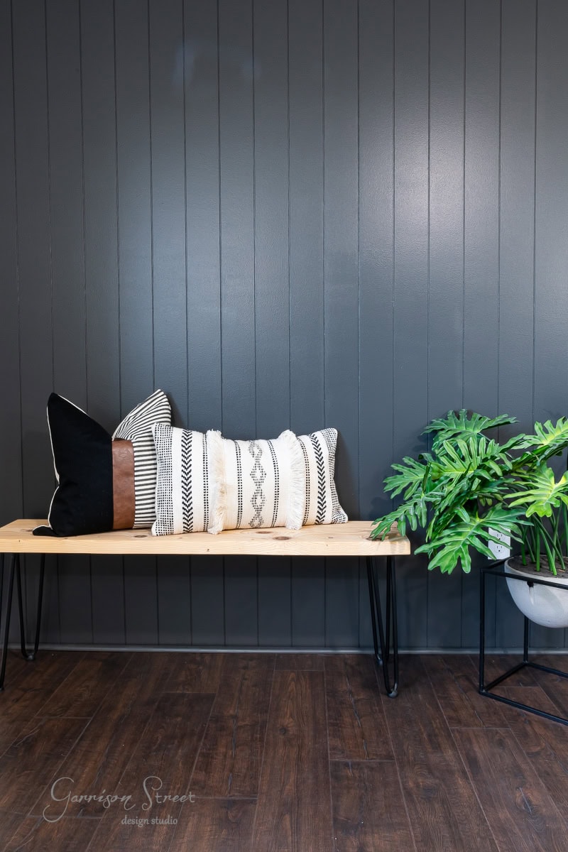 A modern interior featuring a wooden bench with black metal legs, topped with black and white patterned pillows. A potted green plant sits beside the bench, set against a chic DIY shiplap accent wall and rich wooden floor.