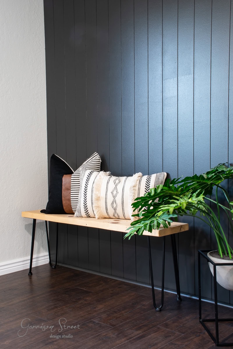 A minimalist room with a wooden bench featuring decorative pillows in various patterns is enhanced by a DIY shiplap accent wall. The bench rests against this stylish backdrop, with a potted plant on one side. The dark wood floor perfectly complements the modern decor.