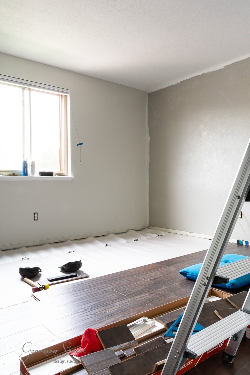 A room under renovation features light gray walls. A window with blinds partially raised lets in natural light, while flooring is being installed. A ladder and tools are scattered in the foreground, alongside a partially laid wooden floor.