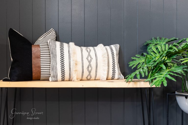 A wooden bench with decorative pillows in various patterns and textures sits against a DIY shiplap accent wall. A potted green plant is placed near the bench, adding a touch of nature to the minimalist design.