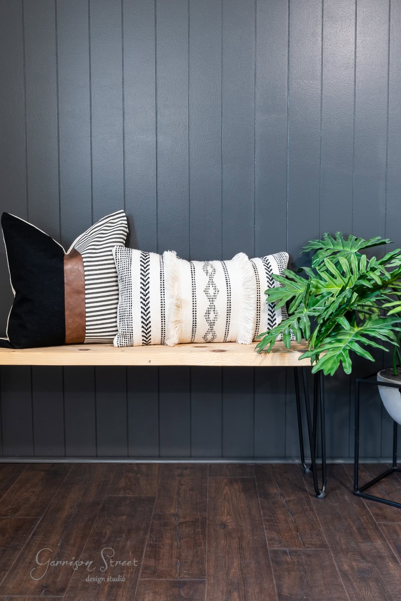 A wooden bench with three decorative pillows featuring black, white, and geometric patterns sits in front of a dark shiplap accent wall. A potted plant with lush green leaves is placed on the right side on a metal stand.