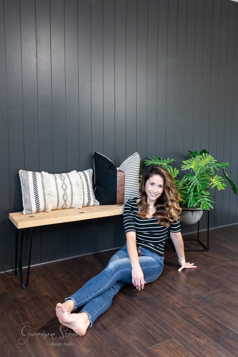 A woman with wavy hair sits on a wooden floor, leaning against a dark DIY shiplap accent wall. She's wearing a striped shirt and jeans, smiling. A wooden bench with decorative pillows and a potted plant are beside her.