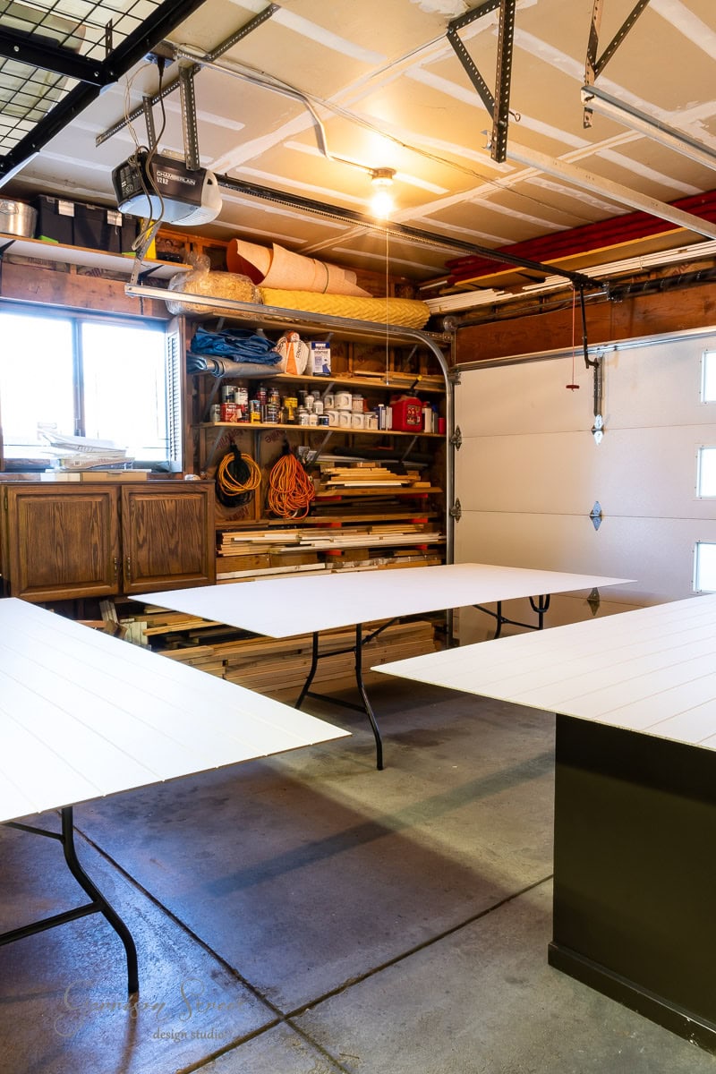 A well-organized garage with tables set up for a DIY project. The walls are lined with shelves holding tools, rolled-up materials, and various supplies. A ceiling light illuminates the workspace, and a garage door is partially visible.