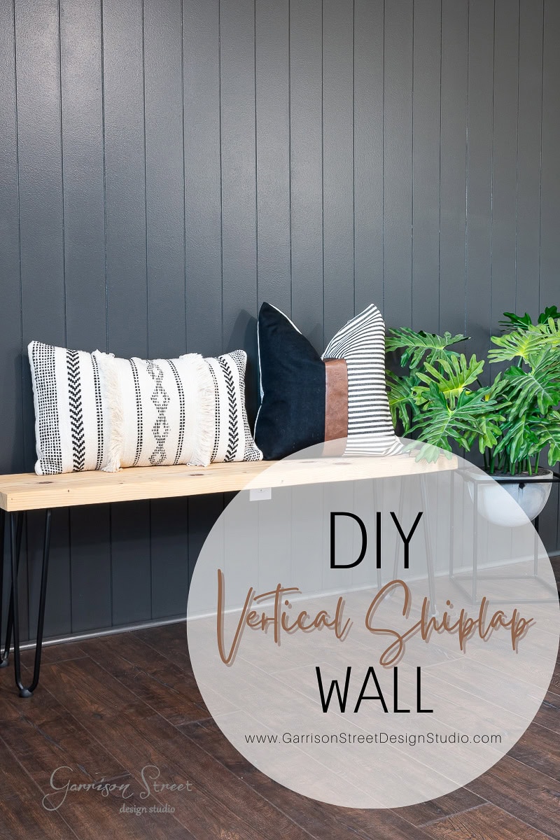 A stylish room features a dark DIY shiplap accent wall, a wooden bench with hairpin legs, decorative pillows, and a plant. The text reads "DIY Vertical Shiplap Wall" with a website link to Garrison Street Design Studio.