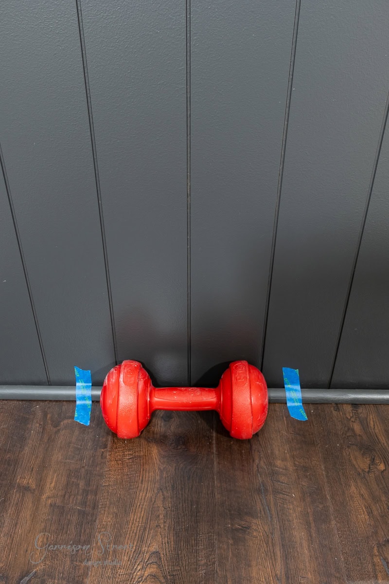 A red dumbbell rests on a wooden floor against a dark gray, DIY shiplap accent wall. Two small pieces of blue tape are affixed to the wall on either side of the dumbbell.