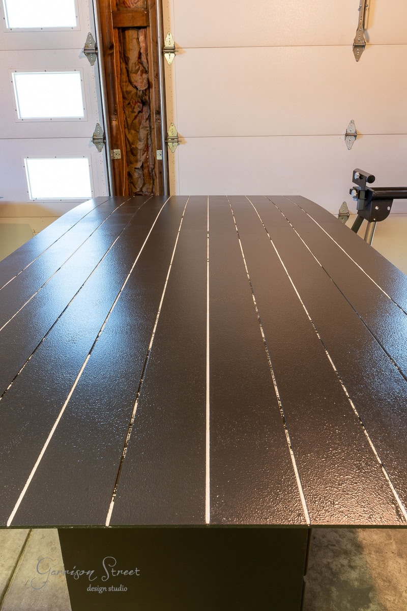 A close-up of a tabletop with a dark surface featuring evenly spaced parallel grooves. The background shows a partially open garage door, revealing a wooden beam and various tools.