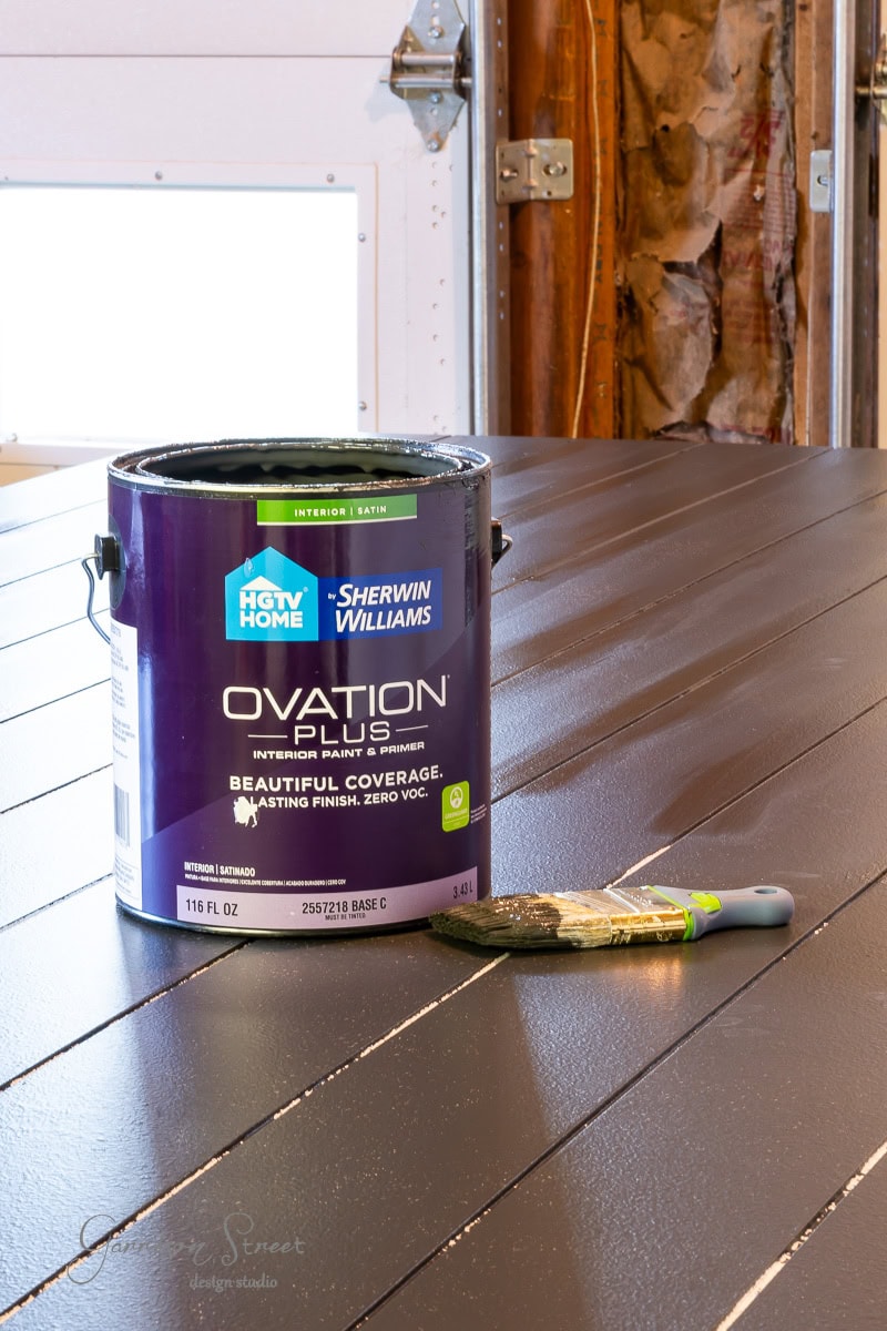 A large purple paint can labeled "HGTV HOME by Sherwin Williams Ovation Plus" sits on a dark, freshly painted wooden surface. A paintbrush lies beside it. The background reveals an unfinished wall with exposed insulation.
