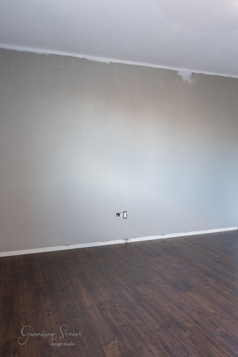 A wall partially painted gray sits above a hardwood floor. Two electrical outlets are visible, surrounded by some unpainted patches. The flooring is dark wood with a smooth, polished finish.