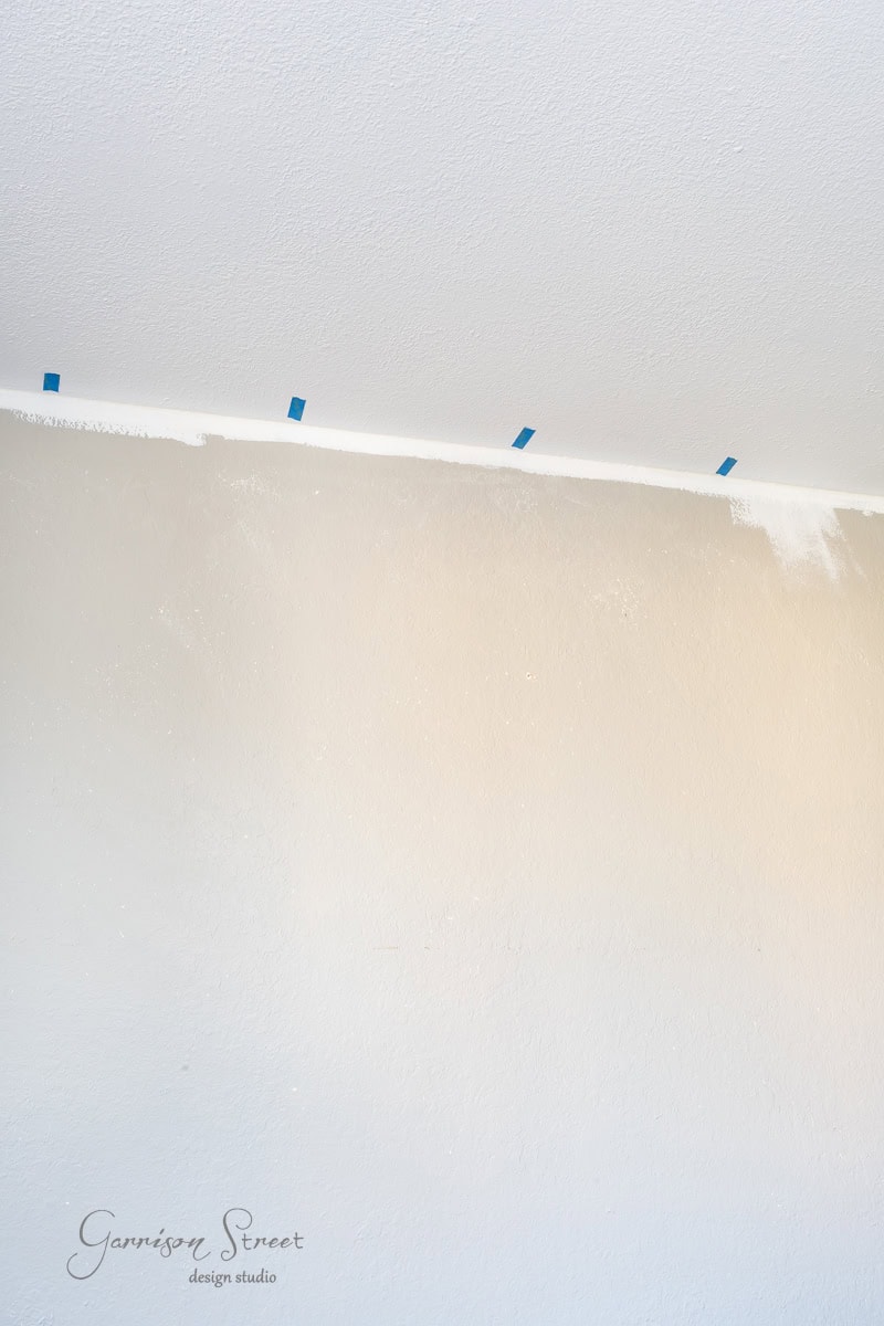 A partially painted wall with white paint on the upper part, taped with small blue pieces for alignment. The paint gradually fades into the previous color. The ceiling is white. Text at the bottom reads "Garrison Street Design Studio."