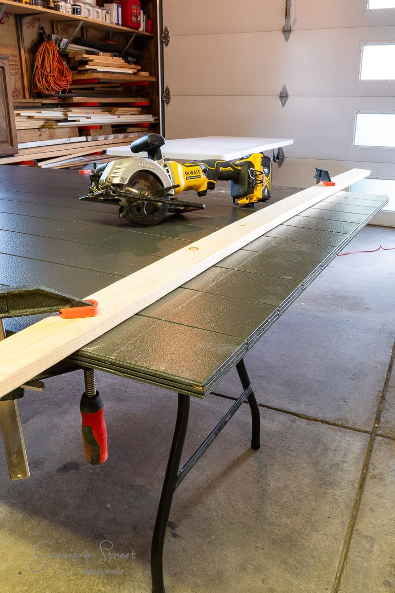 A garage workspace featuring a table saw, clamps, and a wooden plank on a black tabletop. The bright room brims with tools on shelves.