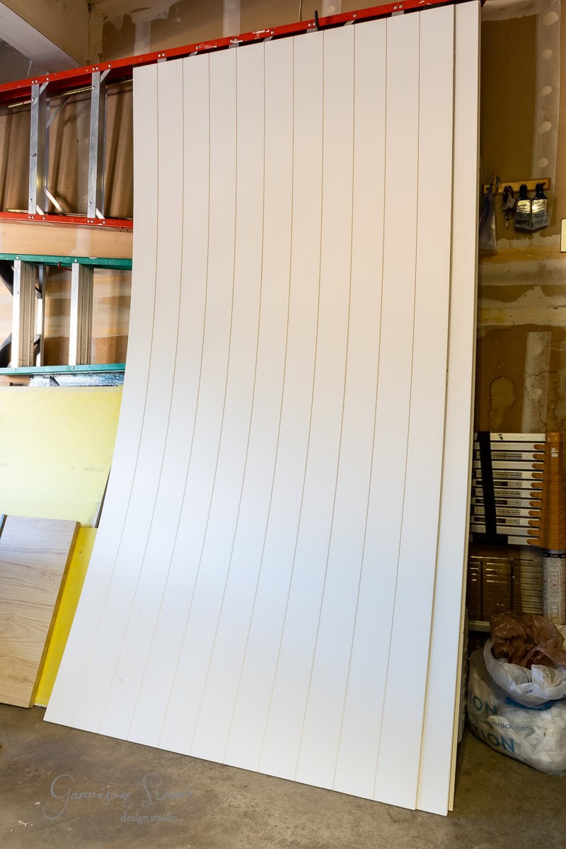 A large, flexible white panel with vertical grooves leans against a wall in a workshop, ready for a DIY closet door makeover. The surrounding area is scattered with construction materials and tools, hinting at the creative transformation about to unfold.