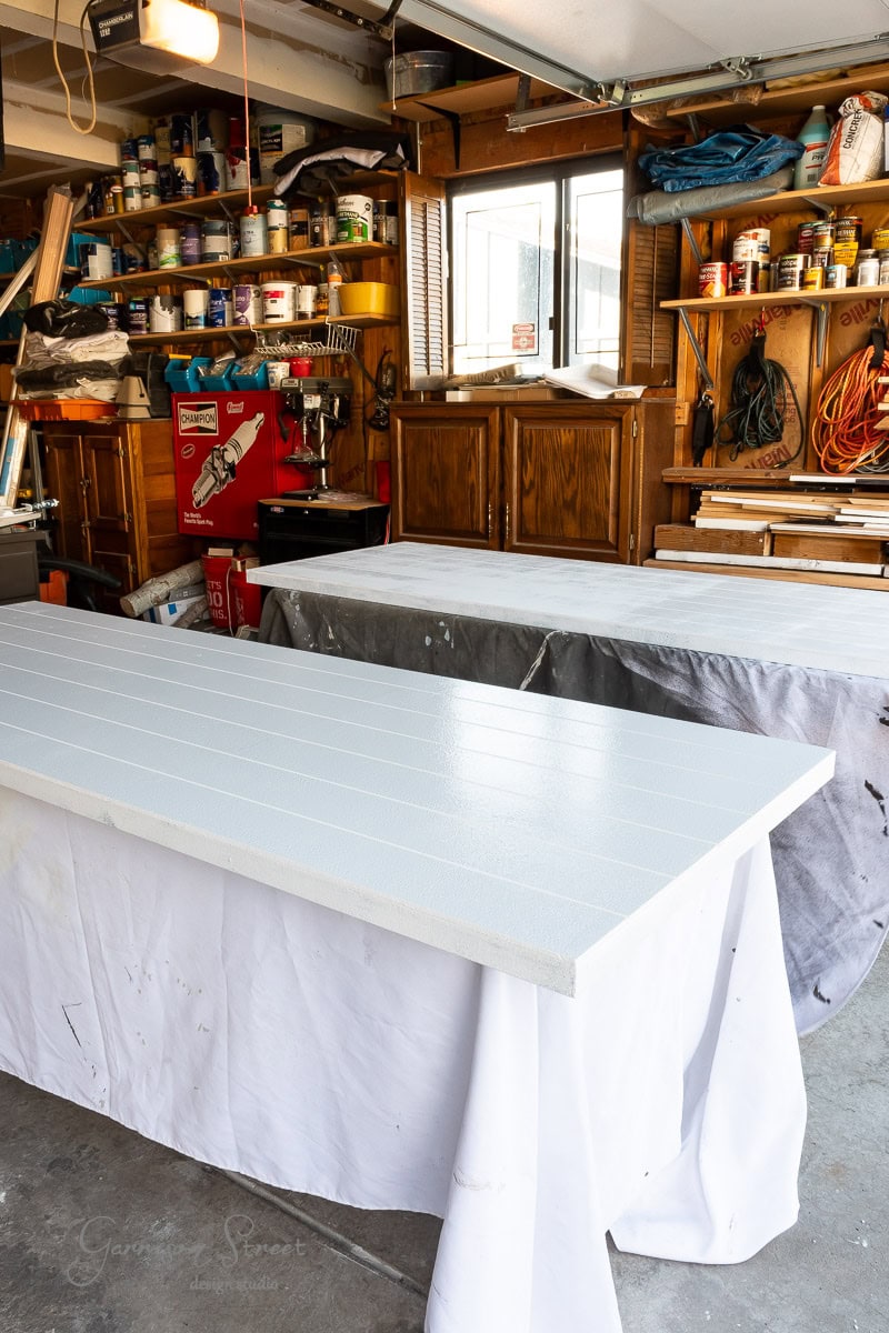 A garage workshop features two long white wooden boards resting on sawhorses with drop cloths, perfect for a DIY closet door makeover. Shelves filled with tools, paint cans, and hardware line the walls, while a window allows natural light into the cluttered space.