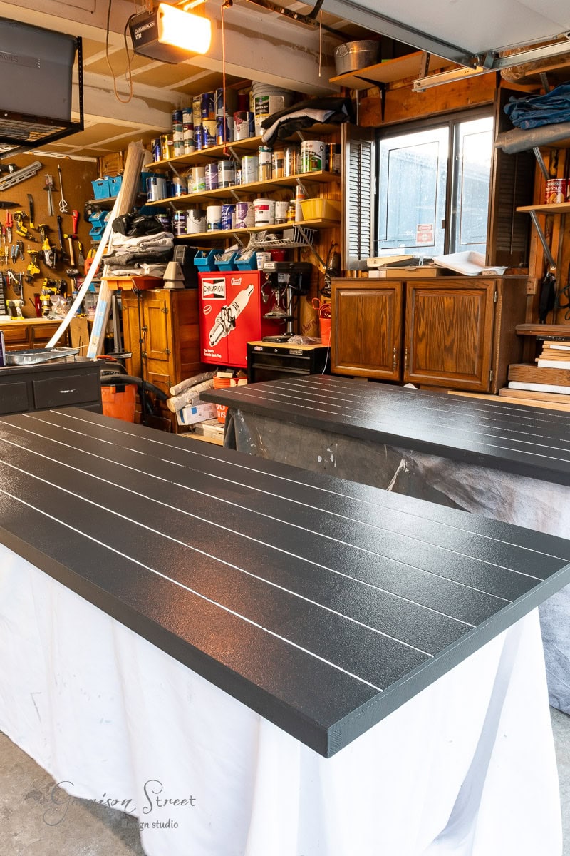 A workshop with two large, dark-colored wooden surfaces, each featuring white lines for a DIY closet door makeover, rests on supports. The background shows shelves filled with various tools, paint cans, and supplies. A window allows light into the cluttered workspace.