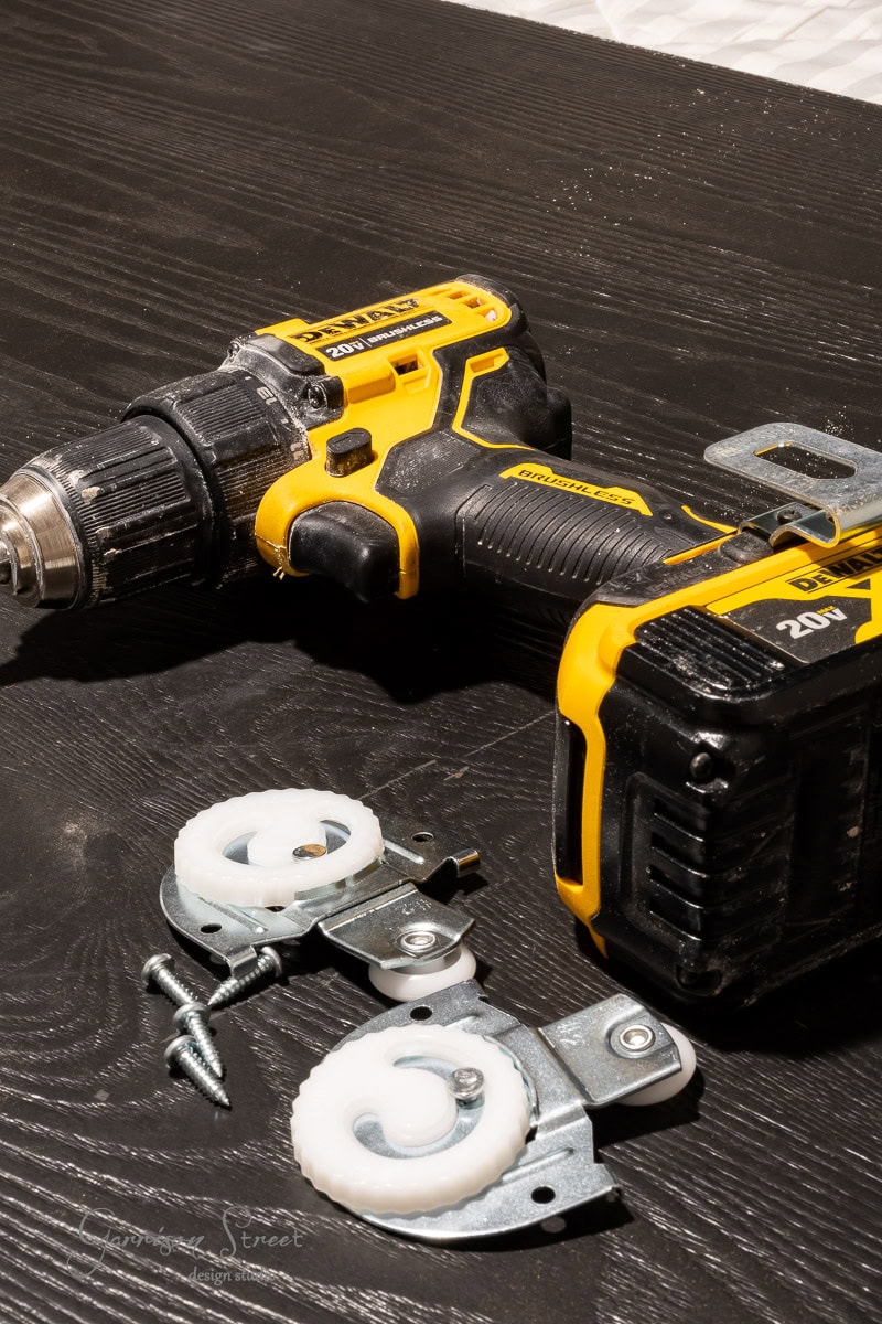 A yellow and black cordless drill is placed on a dark wooden table next to two white roller brackets and screws, perfect for a DIY closet door makeover or installing sliding doors or drawers.