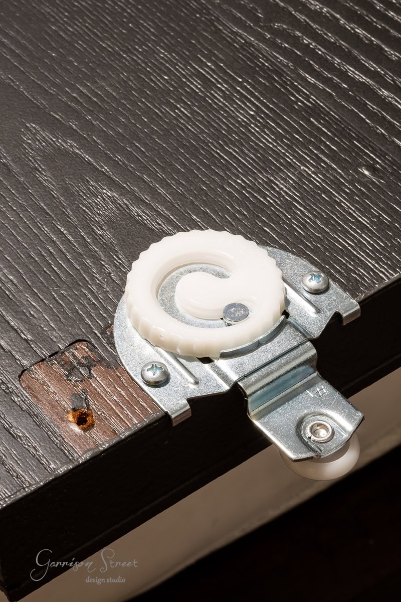 Close-up of a white plastic sliding door roller mechanism, perfect for a DIY closet door makeover, attached to the edge of a black wooden panel with a visible screw and circular hole. The wood grain is evident, and the metal bracket is shiny.