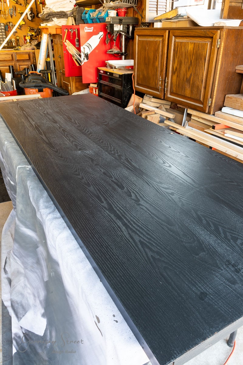 A large wooden table in a workshop, painted black with a glossy finish, serves as the ideal space for your next DIY closet door makeover. Various tools and wooden boards are scattered in the cluttered background, ready to bring creative visions to life.