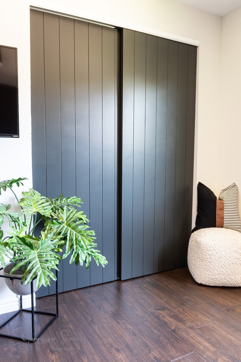 A modern room features dark green vertical panel closet doors, transformed by a stylish DIY closet door makeover. A cozy pouf and pillow rest in the corner, while a potted plant with large leaves adorns the wooden floor. A wall-mounted TV is partially visible, adding to the chic ambiance.