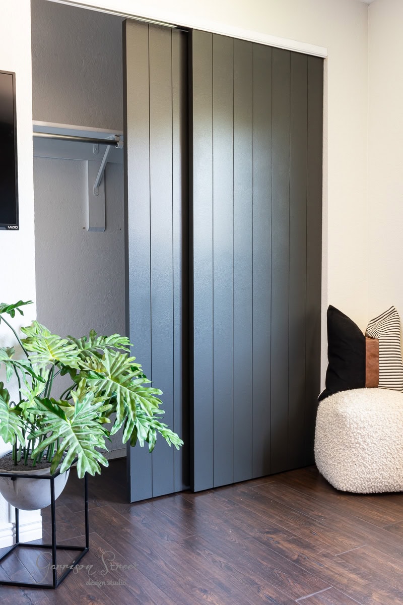 A modern room showcases dark wood flooring and a DIY closet door makeover with a black paneled sliding door, partially open to reveal a stylish interior. A leafy potted plant graces the left, while a cozy round white chair with black and striped pillows completes the scene on the right.