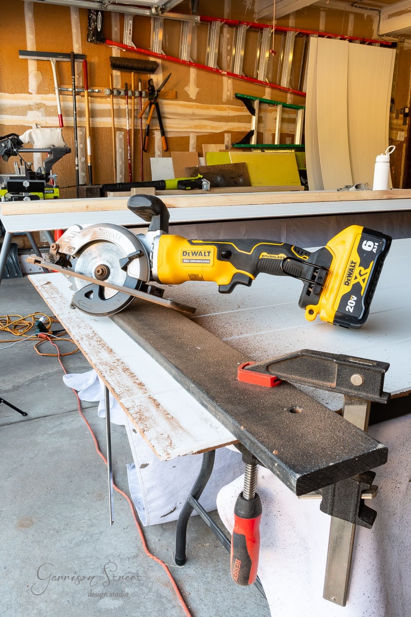 A DeWalt cordless circular saw sits on a piece of metal on a workbench in a garage, ready for action. The background reveals an array of tools and partially constructed wooden panels, hinting at an ongoing DIY closet door makeover.