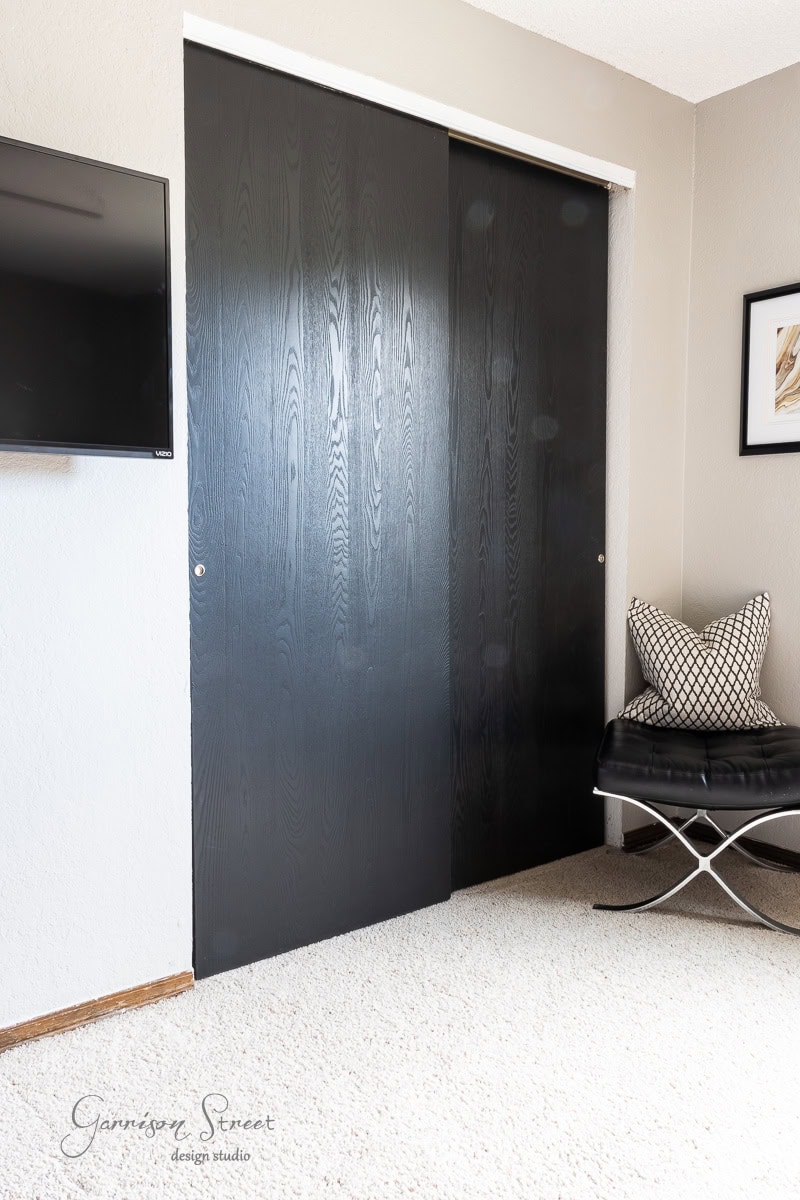 A room features a chic DIY closet door makeover with sliding black wood grain doors, complemented by a wall-mounted TV on the left and a modern chair adorned with decorative cushions on the right. The light beige carpet adds warmth to the stylish space.