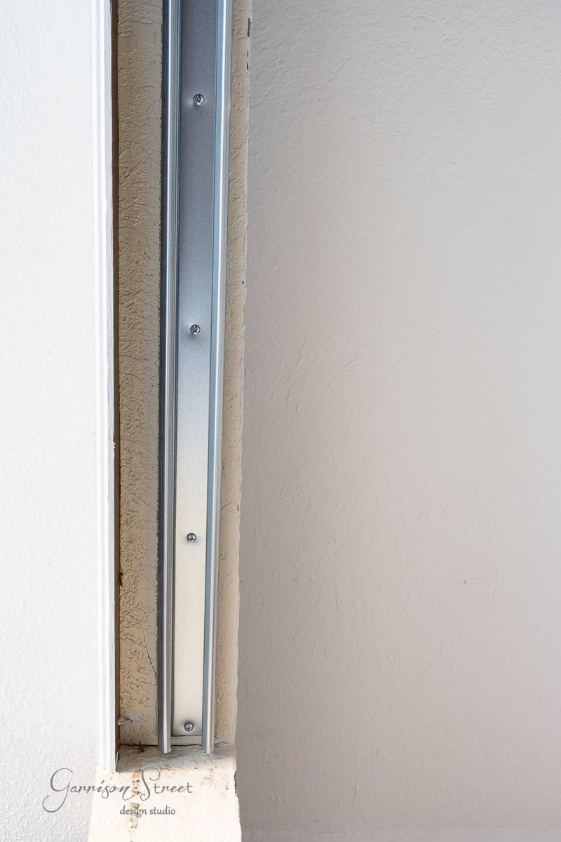 Close-up of a wall corner showcasing a vertical metal bracket attached to the textured white wall, ideal for a DIY closet door makeover. The bracket features evenly spaced screws, and sunlight casts subtle shadows. The bottom left corner displays a faint logo reading "Garrison Street Design Studio.