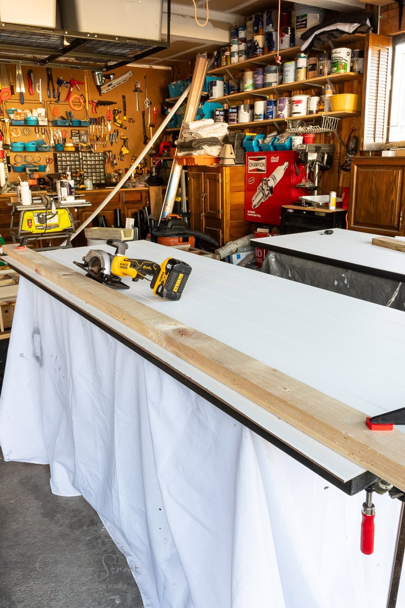 A cluttered workshop filled with tools, paint cans, and a variety of equipment. In the center, a long closet door undergoing a DIY makeover rests on sawhorses, accompanied by power tools and a wooden plank. Shelving and storage are visible in the background.