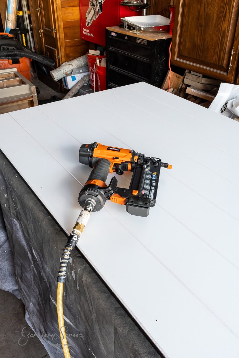 A pneumatic nail gun with an air hose sits on a large white board, ready for your next DIY closet door makeover. In the workshop's background, various tools and equipment promise endless possibilities for transformation.