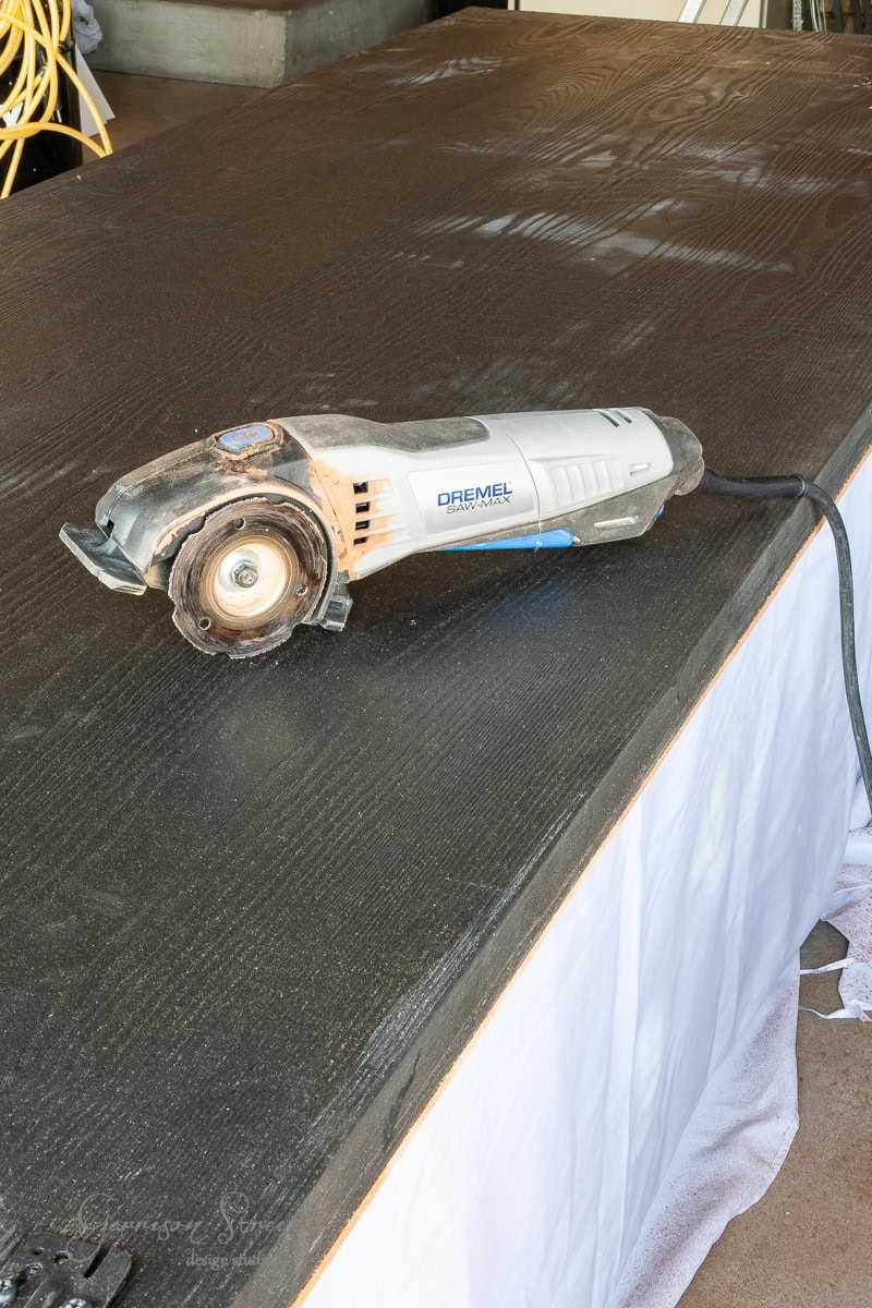 A Dremel power tool sits atop a large dark wooden surface, poised for a DIY closet door makeover. The tool is plugged in, with its cord stretching across the table. Draped cloth and coiled cables provide a fitting backdrop for this craft scene.