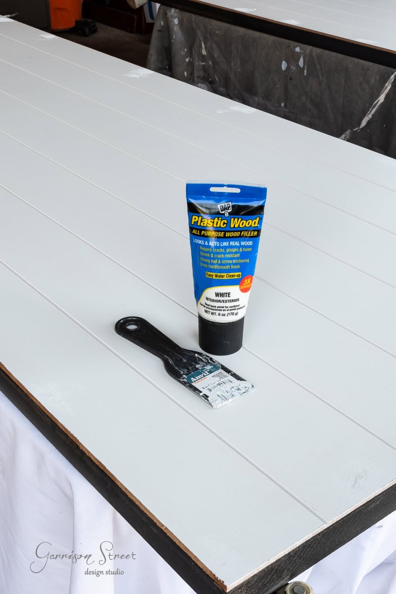 A tube of DAP Plastic Wood rests on a painted wooden panel, perfect for your DIY closet door makeover. Nearby, a paintbrush lies ready to smooth the grooves. In the background, additional painted panels await their transformation.