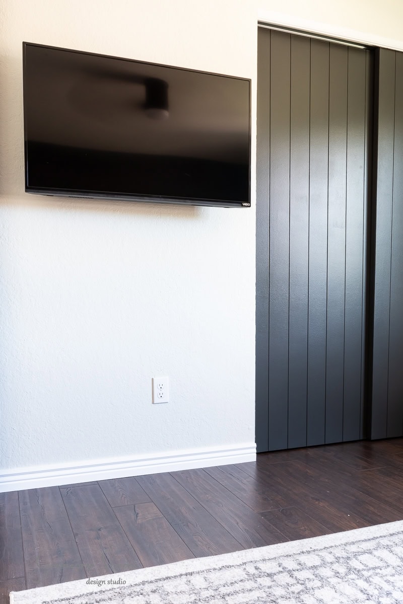 A wall-mounted flat-screen TV is positioned on a white wall adjacent to a modern, dark sliding door. The floor, made of polished dark wood, contrasts beautifully with the room's minimalist design.