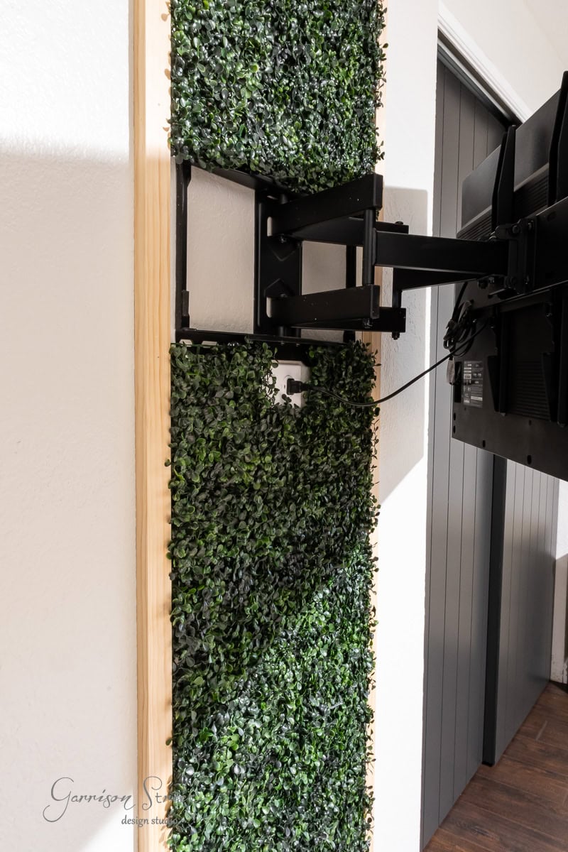A wall-mounted TV on a swivel arm is surrounded by lush green artificial foliage. The foliage elegantly frames the TV and is attached to a wooden panel on the pristine white wall.