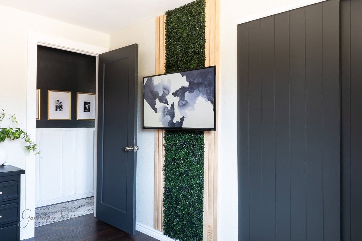Room with dark paneled walls and a door. A framed abstract painting hangs on a DIY green wall of lush greenery. A small nook with framed photos is visible. Dark wood flooring adds warmth to the modern, minimalist design.