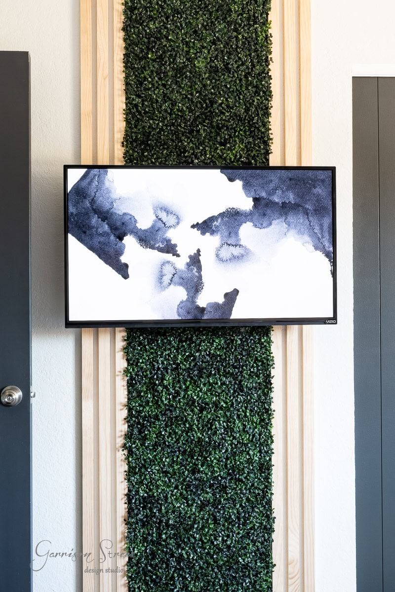 A flat-screen TV showcasing abstract black and white art is mounted on a vertical wooden panel, complemented by a green wall. This stylish setup is nestled between two doors in a room with modern decor.