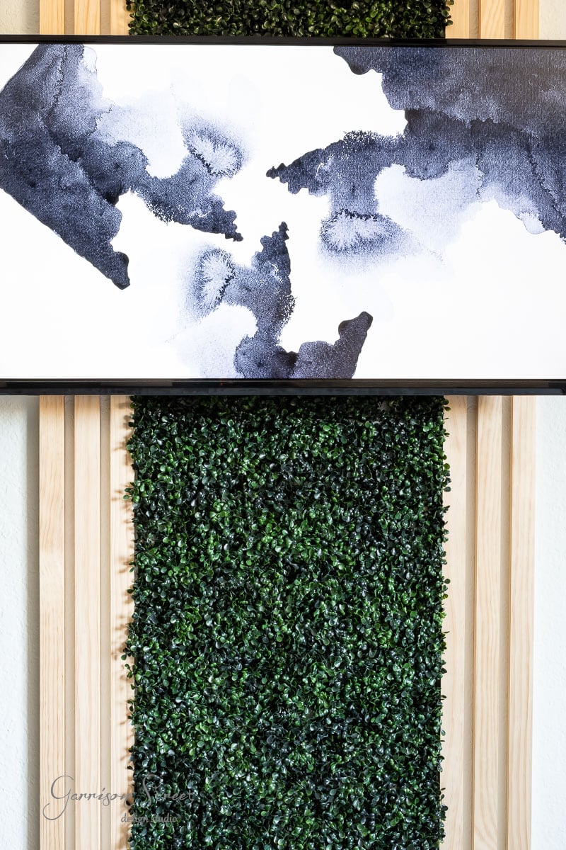 A minimalistic abstract black and white painting is displayed above a vertical panel of lush foliage against the wooden backdrop. The painting showcases organic black ink patterns on a crisp white background.