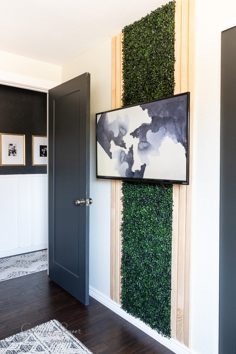 An interior wall showcases a green foliage panel, a modern abstract painting in gray and black shades. A partially open dark gray door reveals an art-adorned hallway. The room features dark wood flooring and white trim.