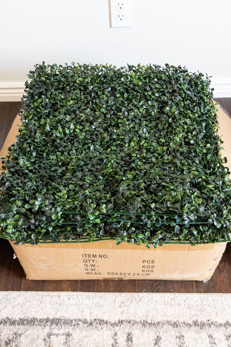A stack of green artificial boxwood hedge panels sits atop a cardboard box on a wooden floor, near a white wall with an electrical outlet visible in the background.