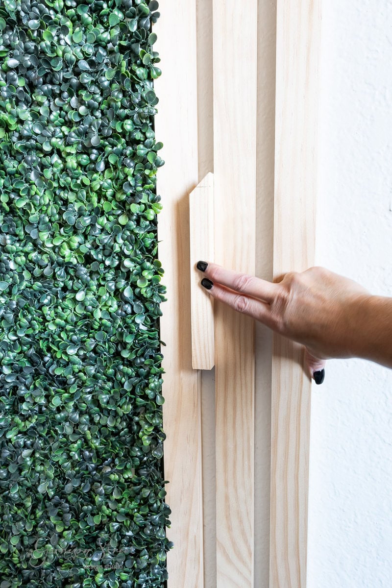 A hand with black nail polish is pulling a wooden lever on a light wooden panel. The panel sits beside a vertical garden brimming with lush green foliage.