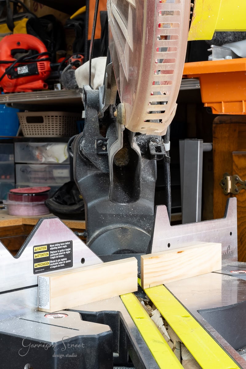 A miter saw is positioned in a workshop, set up to make a precise cut in a piece of wood. Shelves with tools and supplies offer a glimpse into a busy, organized workspace.