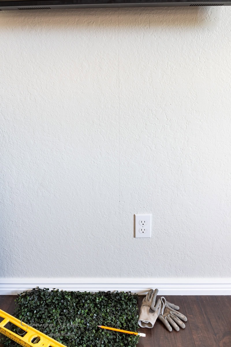 A plain white wall with an electrical outlet. On the floor, gardening gloves, a pencil, a yellow level, and artificial green foliage lie ready. The wooden floor is accented by a white baseboard.