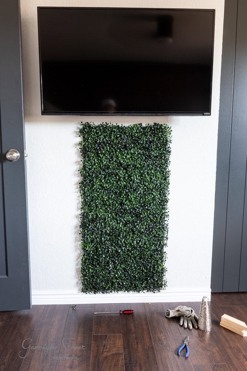 A wall-mounted TV hangs above a small rectangular green wall panel on a white backdrop. Tools like pliers, gloves, and a small red toolbox are scattered across the wooden floor in front.