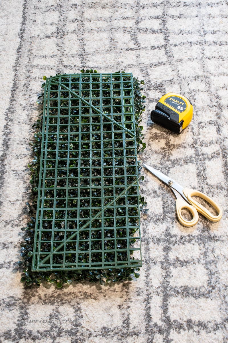 A piece of green artificial grass mat lies on a patterned gray carpet. Nearby are a pair of gold-handled scissors and a yellow tape measure.