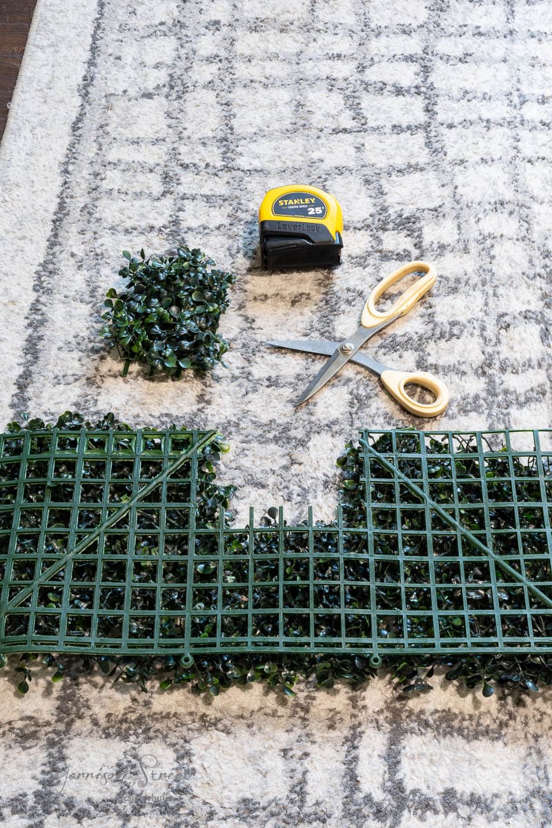 A piece of green lattice is being cut on a patterned rug. Scissors and a yellow tape measure are nearby, alongside a small bunch of leaves.