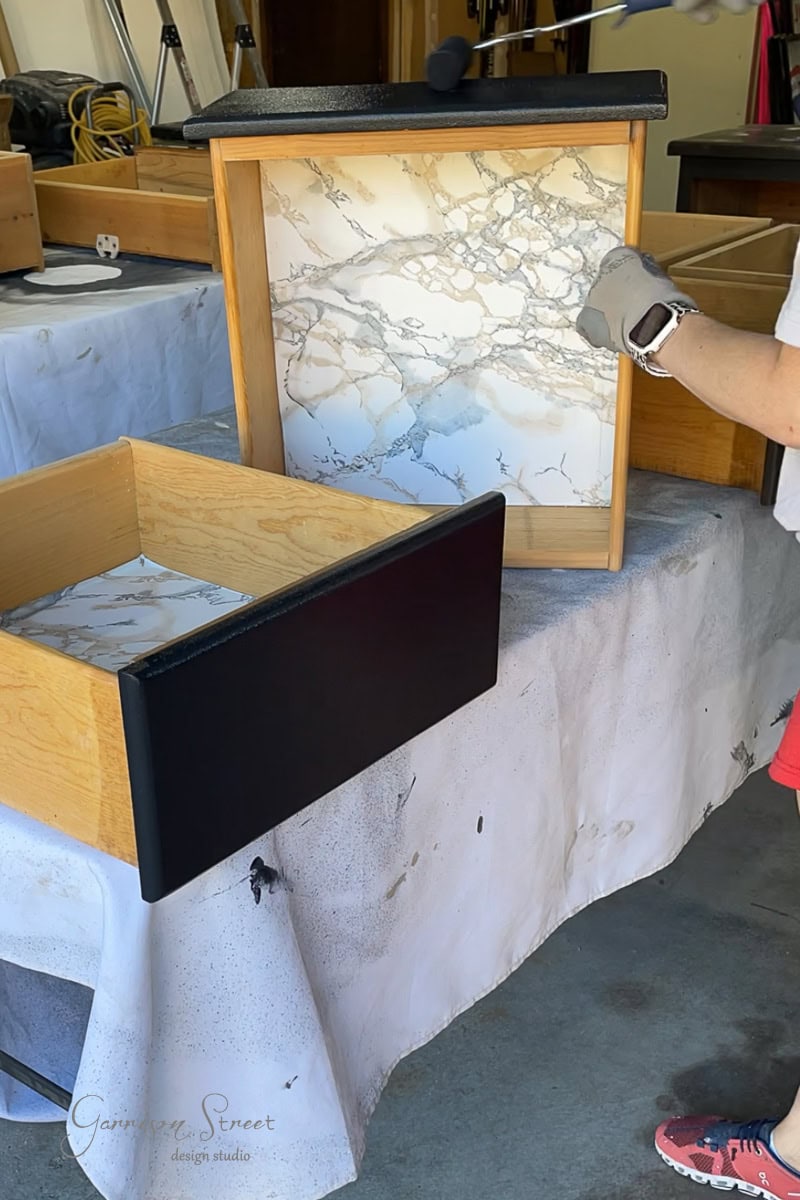 A person wearing a glove holds a wooden drawer with a marble-patterned lining. The drawer features a sleek black front panel and is set in a workshop amidst similar transformed drawers.