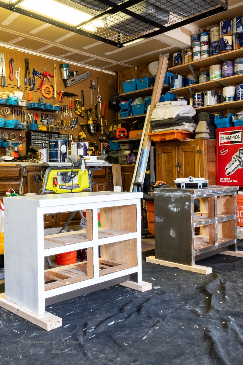 A workshop with shelves full of tools and paint cans. Two partially painted cabinets sit on a tarp, with one painted white and the other dark grey. Various woodworking equipment is visible in the background.