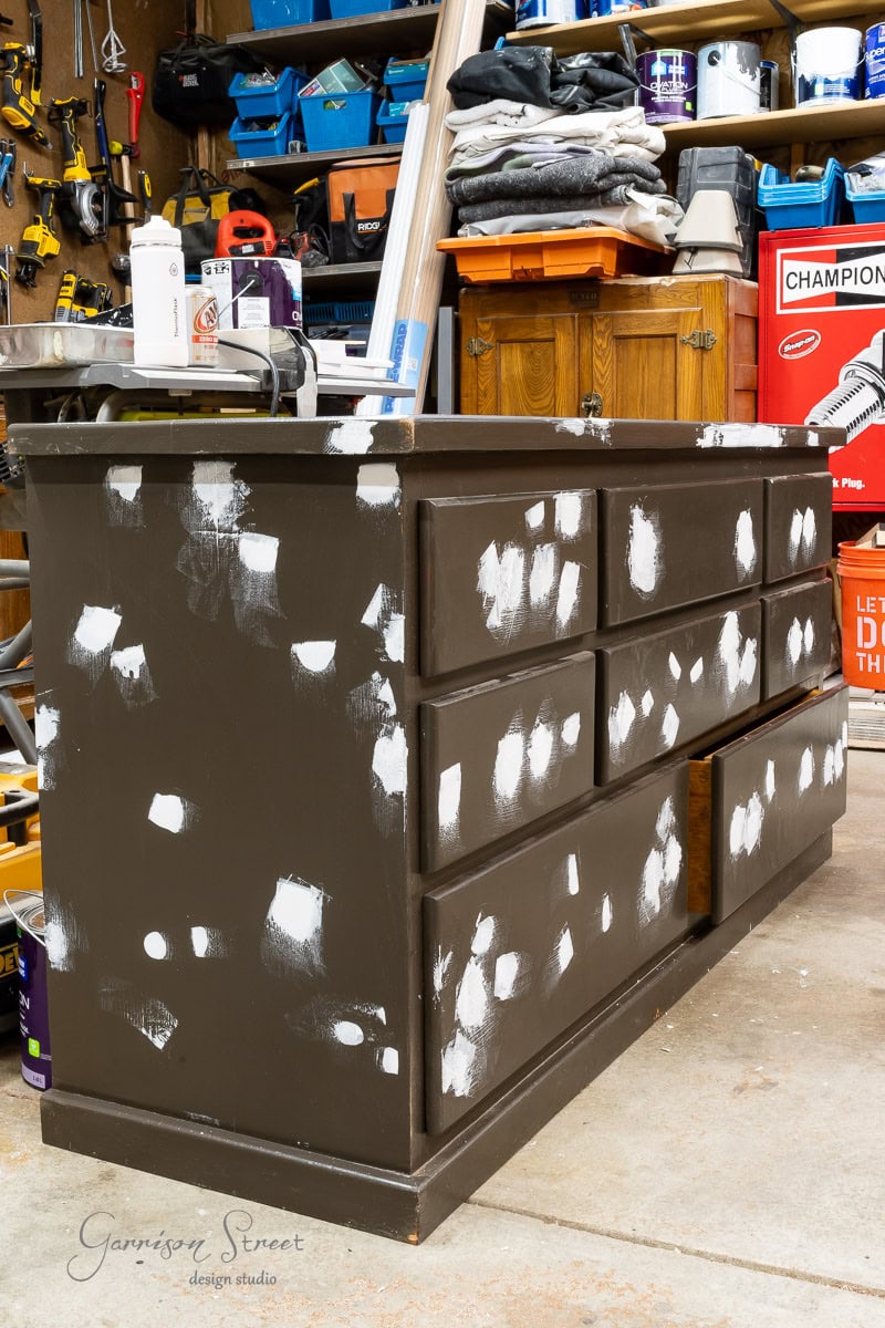 A dark-colored dresser with white patchwork sits in a cluttered garage workshop. Various tools and storage boxes are visible in the background.