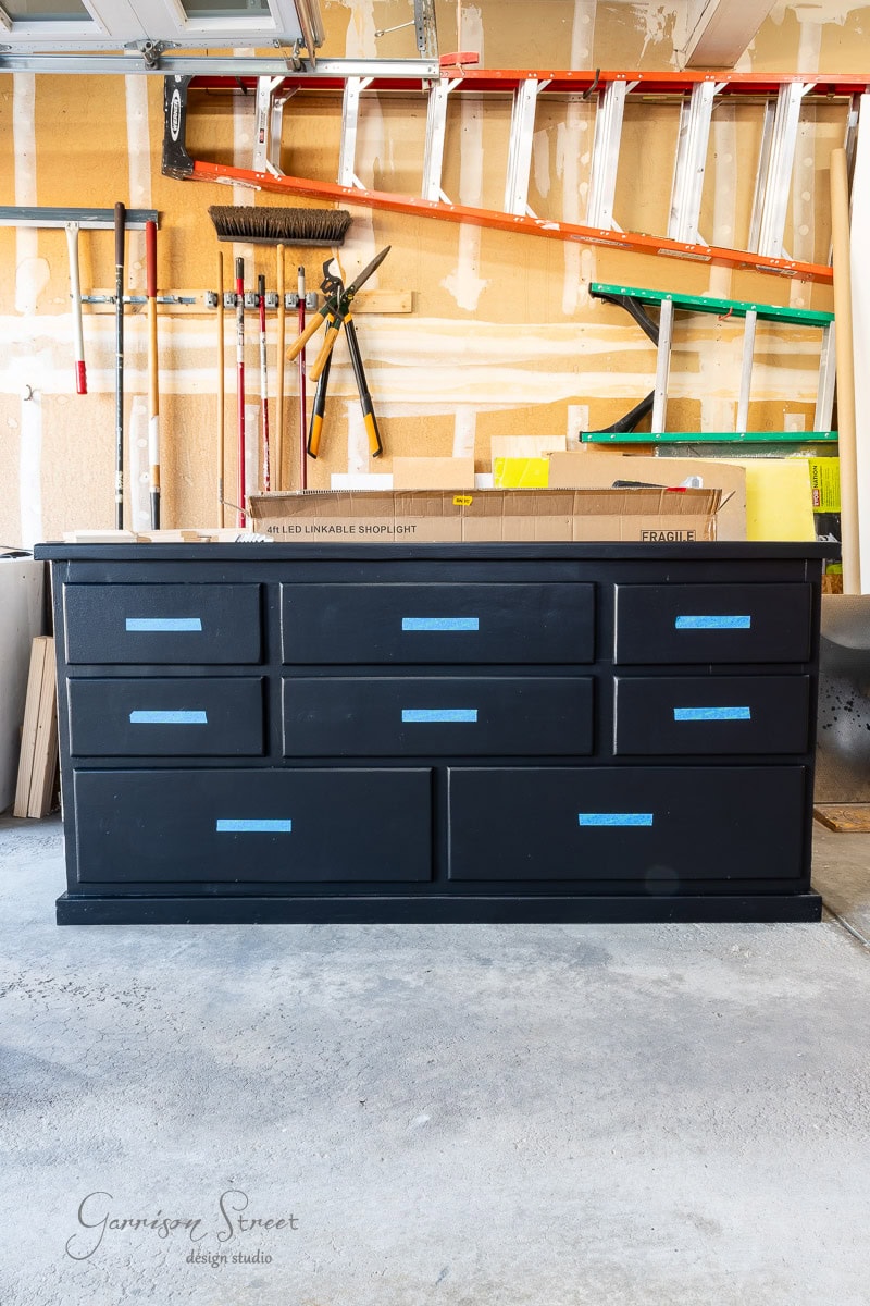 A black dresser stands in a garage workshop with blue tape on each drawer. Surrounding it are various tools, including a broom, rake, and ladders, set against a beige wall showcasing visible drywall.