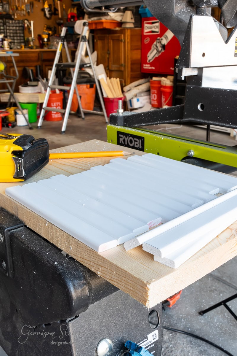 A woodworking shop featuring a miter saw with white trim pieces on a wooden workbench. In the background, various tools and equipment, including buckets and a toolbox, complete this creative space.