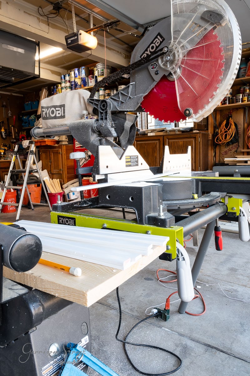 A Ryobi miter saw is set up in a garage workshop, positioned on a stand with a wooden plank. The garage brims with various tools and equipment neatly arranged on shelves and hanging on the walls.