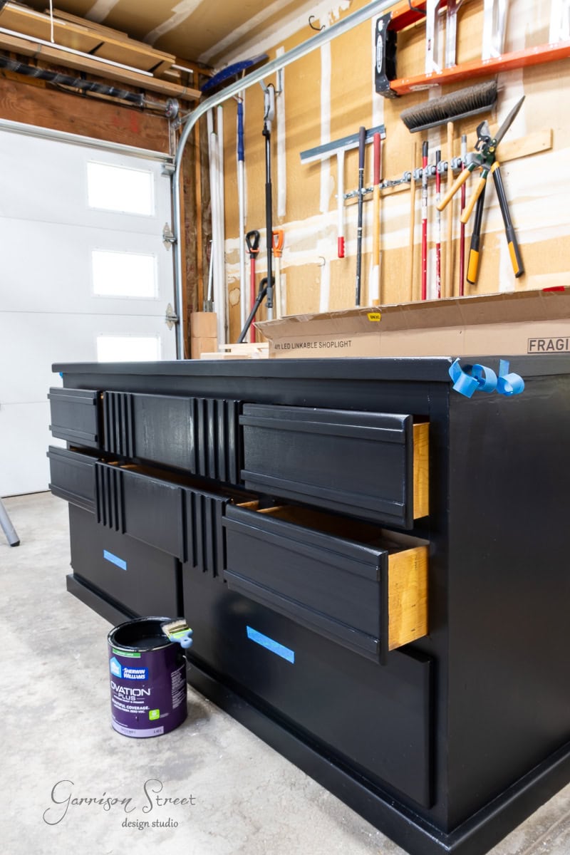 A black-painted dresser stands in the garage with its drawers slightly open. Tools hang on the wall in the background, while a can of dark paint rests nearby on the floor.