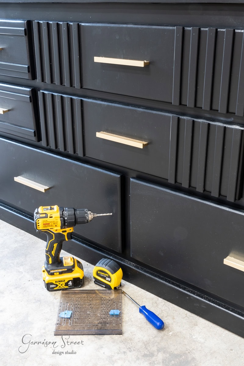 A black dresser with modern gold handles sits on a light floor. In front of the dresser, you'll find a yellow power drill, a yellow tape measure, a blue-handled screwdriver, and a metal ruler.